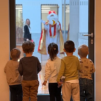 5 Kleinkinder in der Mukki Kinderbetreuung sehen bei einer Glastür hinaus vor der der Nikolaus steht und den Kindern winkt.