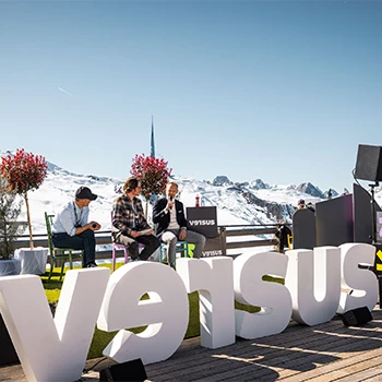 Outdoor Podiumsdiskussion beim versus Festival mit Schriftzug "versus" im Vordergrund und verschneiten Bergen im Hintergrund